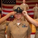 A U.S. Navy promotion ceremony is taking place in front of an American flag. Two senior enlisted sailors are placing a Chief Petty Officer combination cover (hat) on a newly promoted sailor, a traditional ritual in Navy CPO pinning/frocking ceremonies. The recipient is smiling, indicating this is a celebratory milestone event.