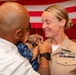 A military promotion or pinning ceremony is taking place, with a civilian man pinning a new insignia or rank device onto a female U.S. Navy officer's uniform collar. The officer is smiling and wearing a tan/khaki Navy service uniform adorned with multiple ribbons and a warfare qualification badge. An American flag is displayed in the background, consistent with a formal military ceremony setting.