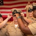 A U.S. Navy promotion or pinning ceremony is taking place, with two senior enlisted personnel placing a Chief Petty Officer cover (hat) on a newly promoted sailor. The ceremony is conducted in front of a large American flag, consistent with formal military advancement traditions. Multiple uniformed Navy personnel are present, some wearing service ribbons and Chief Petty Officer insignia.