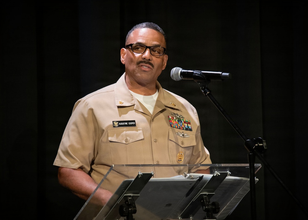 A U.S. Navy officer in Service Khaki uniform is speaking at a clear acrylic podium with a microphone. His name tag reads 'Augustine Cooper' and he displays multiple service ribbons and medals on his uniform. The setting appears to be a formal military ceremony or official speaking engagement against a dark background.