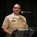 A U.S. Navy officer in Service Khaki uniform is speaking at a clear acrylic podium with a microphone. His name tag reads 'Augustine Cooper' and he displays multiple service ribbons and medals on his uniform. The setting appears to be a formal military ceremony or official speaking engagement against a dark background.