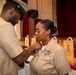 Two U.S. Navy personnel in tan service uniforms are engaged in a pinning ceremony, with one service member attaching an insignia or device to the collar of the other. The setting appears to be a formal military ceremony in an auditorium or hall, with red curtains and wooden gift bags visible in the background. The recipient appears emotional, suggesting this is a significant promotion or advancement milestone.