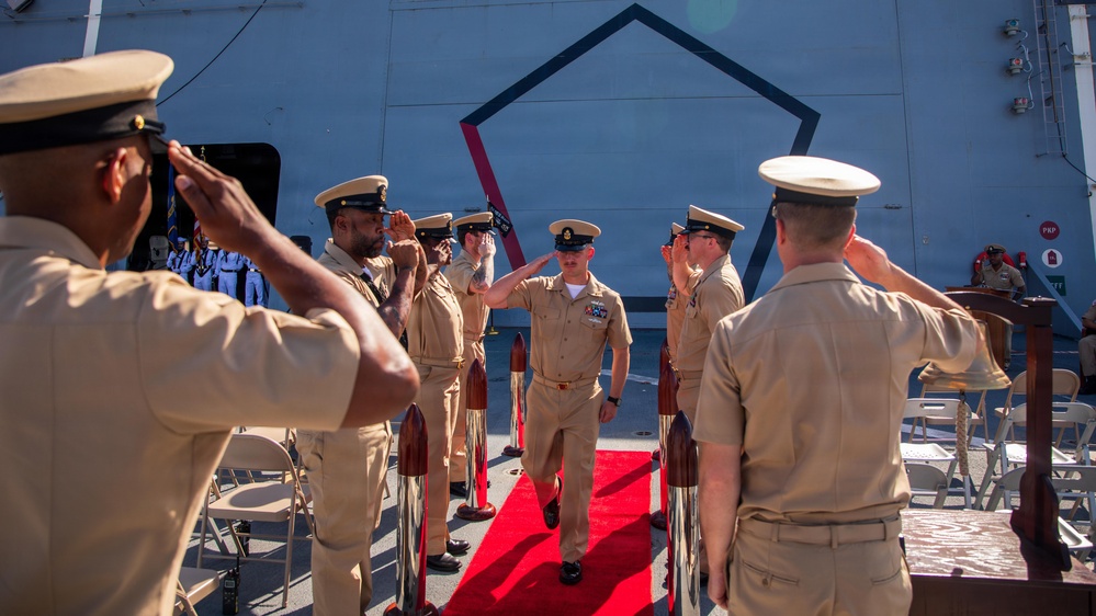 A formal U.S. Navy military ceremony is taking place on the deck of a naval vessel, with personnel in khaki service uniforms rendering salutes to an officer walking along a red carpet. The scene appears to be a change of command or similar official ceremony, indicated by the red carpet, ceremonial stanchions, and rows of chairs visible in the background. Multiple sailors are lined up on both sides forming an honor corridor.
