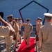 A formal U.S. Navy military ceremony is taking place on the deck of a naval vessel, with personnel in khaki service uniforms rendering salutes to an officer walking along a red carpet. The scene appears to be a change of command or similar official ceremony, indicated by the red carpet, ceremonial stanchions, and rows of chairs visible in the background. Multiple sailors are lined up on both sides forming an honor corridor.