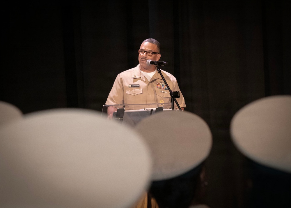 A U.S. Navy officer in tan service uniform (Service Khaki) stands at a podium with a microphone, appearing to deliver a speech or address. The foreground shows blurred white Navy covers (hats) of seated audience members, suggesting a formal military ceremony or event. The dark stage backdrop and lighting focus attention on the speaker.