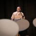 A U.S. Navy officer in tan service uniform (Service Khaki) stands at a podium with a microphone, appearing to deliver a speech or address. The foreground shows blurred white Navy covers (hats) of seated audience members, suggesting a formal military ceremony or event. The dark stage backdrop and lighting focus attention on the speaker.