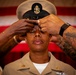 A U.S. Navy Chief Petty Officer promotion ceremony is taking place, with two individuals placing the distinctive khaki combination cover (cap) onto the head of the newly promoted Chief. The subject is wearing the khaki service uniform with Chief Petty Officer collar devices and a warfare qualification badge. An American flag is visible in the background, consistent with a formal military ceremony setting.