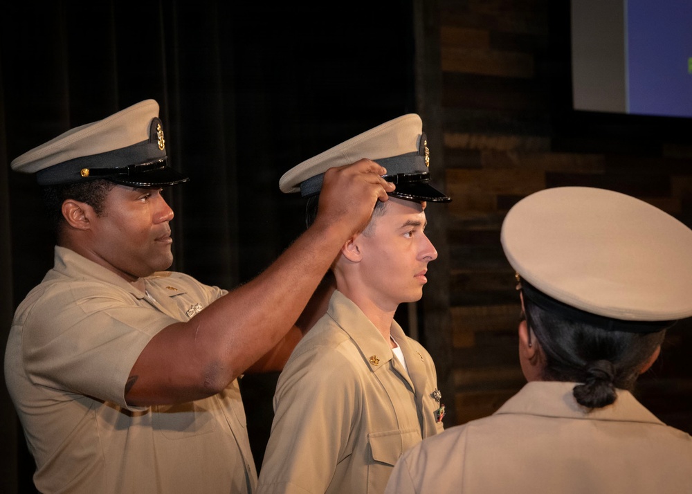 A military promotion or advancement ceremony is taking place, with one uniformed service member placing a cover (hat) on another service member's head. All individuals are wearing tan/khaki uniforms consistent with U.S. Navy service dress uniforms. A third uniformed individual is visible in the foreground with their back to the camera.