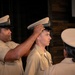 A military promotion or advancement ceremony is taking place, with one uniformed service member placing a cover (hat) on another service member's head. All individuals are wearing tan/khaki uniforms consistent with U.S. Navy service dress uniforms. A third uniformed individual is visible in the foreground with their back to the camera.