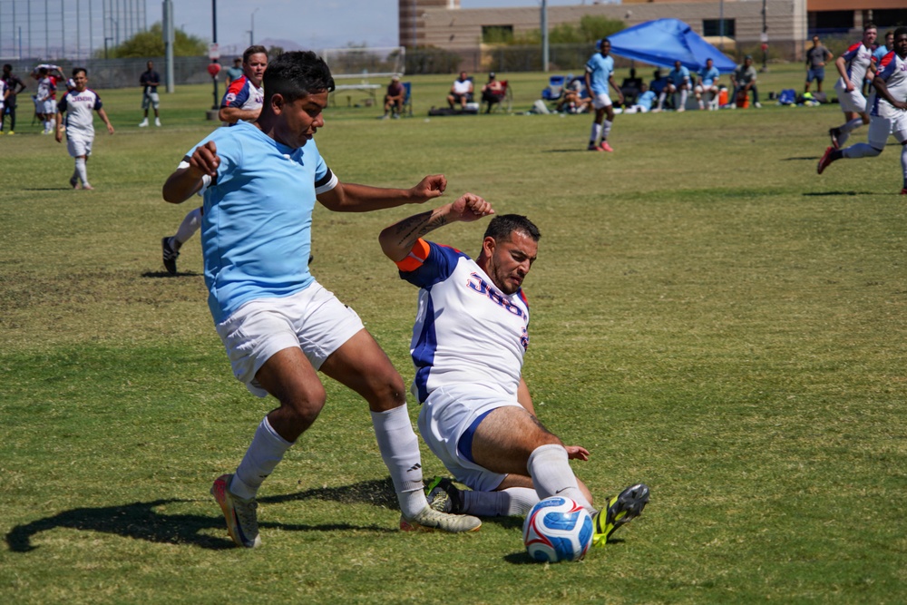 An outdoor amateur or semi-professional soccer match is in progress on a grass field under bright sunlight. Two players in the foreground are engaged in a slide tackle challenge for the ball, with one player in a light blue jersey and white shorts and another in a white jersey executing the slide. Spectators, additional players, and a blue canopy tent are visible in the background.