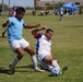 An outdoor amateur or semi-professional soccer match is in progress on a grass field under bright sunlight. Two players in the foreground are engaged in a slide tackle challenge for the ball, with one player in a light blue jersey and white shorts and another in a white jersey executing the slide. Spectators, additional players, and a blue canopy tent are visible in the background.