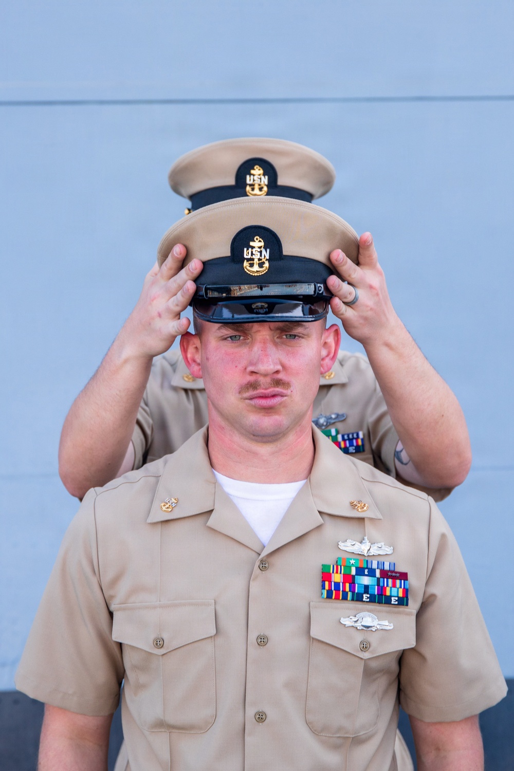 A U.S. Navy Chief Petty Officer is having his combination cover (cap) placed on his head by another person standing behind him during what appears to be a Chief Petty Officer pinning or frocking ceremony. The foreground sailor wears khaki service uniform adorned with multiple ribbons, warfare devices, and collar insignia. This is a traditional Navy milestone ceremony marking advancement to the Chief Petty Officer ranks (E-7 through E-9).
