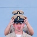 A U.S. Navy Chief Petty Officer is having his combination cover (cap) placed on his head by another person standing behind him during what appears to be a Chief Petty Officer pinning or frocking ceremony. The foreground sailor wears khaki service uniform adorned with multiple ribbons, warfare devices, and collar insignia. This is a traditional Navy milestone ceremony marking advancement to the Chief Petty Officer ranks (E-7 through E-9).