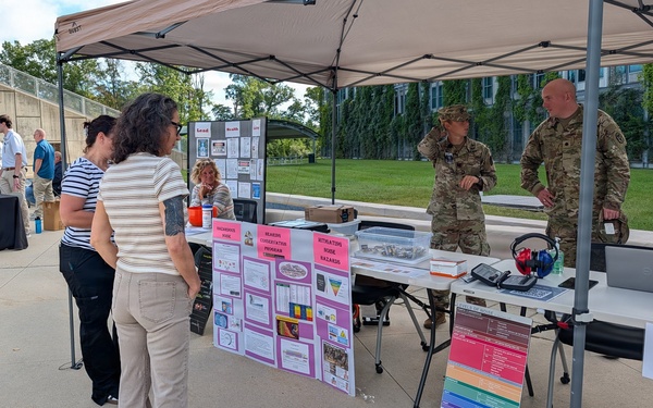 An outdoor health and safety information booth is set up under a tan canopy tent, staffed by two military personnel in camouflage uniforms. The booth focuses on hearing conservation and noise hazard awareness, with display boards and equipment visible. Two civilian visitors are engaging with the exhibit while a third civilian sits behind the table.