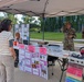 An outdoor health and safety information booth is set up under a tan canopy tent, staffed by two military personnel in camouflage uniforms. The booth focuses on hearing conservation and noise hazard awareness, with display boards and equipment visible. Two civilian visitors are engaging with the exhibit while a third civilian sits behind the table.