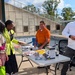 Four people are gathered around a folding table at an outdoor event, likely a health fair or informational booth. Two women on the left are reviewing materials on the table while a man in an orange polo shirt stands behind it, and another man in a white long-sleeve shirt stands to the right. The setting appears to be a plaza or courtyard with a concrete memorial wall featuring engraved names in the background.