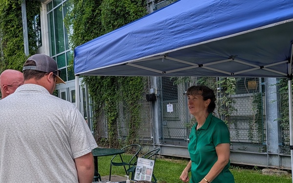 An outdoor educational or vendor booth event is taking place alongside a building covered in ivy. A woman in a green polo shirt stands behind a table draped with animal pelts/furs, speaking with two men. The setting appears to be a public fair or nature/wildlife education event.