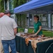 An outdoor educational or vendor booth event is taking place alongside a building covered in ivy. A woman in a green polo shirt stands behind a table draped with animal pelts/furs, speaking with two men. The setting appears to be a public fair or nature/wildlife education event.
