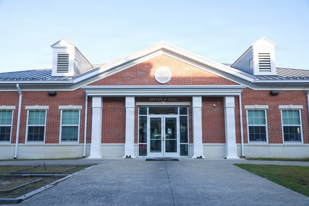 The image shows the front exterior of a brick building identified as 'Dwyer Hall.' The building features a classical architectural style with white columns, a gabled roofline, and a metal roof with cupolas on either side. The entrance is framed by a portico with double glass doors and a paved walkway leading up to it.