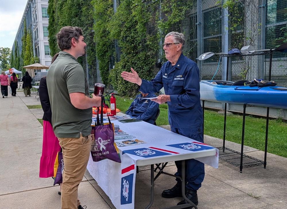 An outdoor community event or fair where a U.S. Coast Guard Auxiliary member in blue uniform is speaking with a civilian visitor at an informational booth. The table is covered with a Coast Guard-branded tablecloth and displays various materials, with a blue kayak visible on a rack nearby. The setting appears to be along a walkway beside a building covered in ivy vegetation.