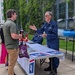 An outdoor community event or fair where a U.S. Coast Guard Auxiliary member in blue uniform is speaking with a civilian visitor at an informational booth. The table is covered with a Coast Guard-branded tablecloth and displays various materials, with a blue kayak visible on a rack nearby. The setting appears to be along a walkway beside a building covered in ivy vegetation.