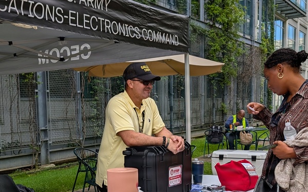 An outdoor event booth for the U.S. Army Communications-Electronics Command (CECOM) featuring a Stop the Bleed or first aid training display. Two booth staff members interact with a visitor at a table covered with medical training mannequin limbs, tourniquets, and first aid supplies. The event appears to be a public outreach or safety awareness fair held in an urban plaza setting.