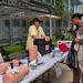 An outdoor event booth for the U.S. Army Communications-Electronics Command (CECOM) featuring a Stop the Bleed or first aid training display. Two booth staff members interact with a visitor at a table covered with medical training mannequin limbs, tourniquets, and first aid supplies. The event appears to be a public outreach or safety awareness fair held in an urban plaza setting.