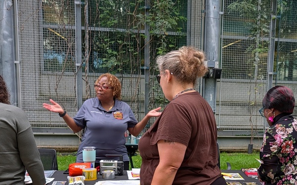An outdoor informational booth event where a presenter in a gray uniform shirt is speaking to attendees gathered around a black-draped table. The booth displays NOAA/National Weather Service materials, including a flood safety sign with a warning triangle. The setting appears to be a courtyard or outdoor area with a glass and metal structure in the background.