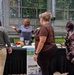 An outdoor informational booth event where a presenter in a gray uniform shirt is speaking to attendees gathered around a black-draped table. The booth displays NOAA/National Weather Service materials, including a flood safety sign with a warning triangle. The setting appears to be a courtyard or outdoor area with a glass and metal structure in the background.