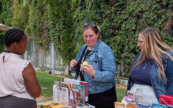 An outdoor informational booth staffed by two women is engaging with a visitor at what appears to be a community health or family services event. The table is covered with a green and yellow tablecloth reading 'Families Are Essential to Readiness' and is stocked with brochures, pamphlets, and promotional materials. One staff member is demonstrating or explaining a small item to the visitor.