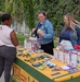 An outdoor informational booth staffed by two women is engaging with a visitor at what appears to be a community health or family services event. The table is covered with a green and yellow tablecloth reading 'Families Are Essential to Readiness' and is stocked with brochures, pamphlets, and promotional materials. One staff member is demonstrating or explaining a small item to the visitor.