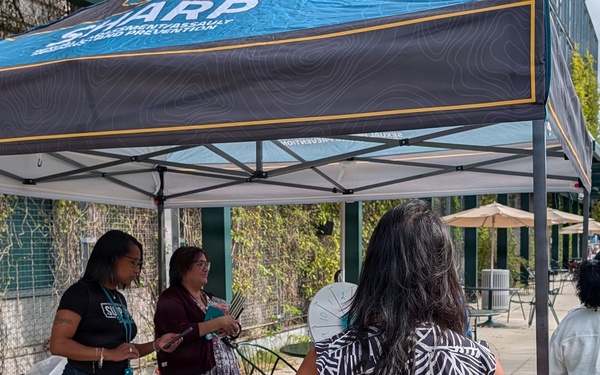 An outdoor event or fair featuring a SHARP (Sexual Harassment/Assault Response and Prevention) program booth under a teal and black canopy tent. Several people are gathered around a table with a teal tablecloth, with booth representatives speaking to attendees. The setting appears to be a paved outdoor area near a building, likely on a military installation or campus.