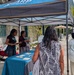 An outdoor event or fair featuring a SHARP (Sexual Harassment/Assault Response and Prevention) program booth under a teal and black canopy tent. Several people are gathered around a table with a teal tablecloth, with booth representatives speaking to attendees. The setting appears to be a paved outdoor area near a building, likely on a military installation or campus.