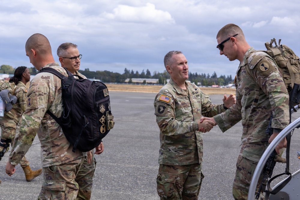 A group of U.S. Army soldiers in OCP (Operational Camouflage Pattern) uniforms are gathered on what appears to be a military airfield tarmac. A senior officer is shaking hands with a soldier carrying a tactical backpack, suggesting a greeting or farewell ceremony. Additional soldiers and personnel are visible in the background near what appears to be a flight line.