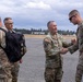 A group of U.S. Army soldiers in OCP (Operational Camouflage Pattern) uniforms are gathered on what appears to be a military airfield tarmac. A senior officer is shaking hands with a soldier carrying a tactical backpack, suggesting a greeting or farewell ceremony. Additional soldiers and personnel are visible in the background near what appears to be a flight line.