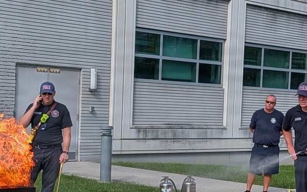 A fire safety training demonstration is taking place outdoors near an industrial or municipal building. A civilian in a blazer and khaki pants is being guided by a firefighter instructor on how to use a fire extinguisher on a controlled propane-fueled training fire. Several firefighters and onlookers are present, observing the exercise.