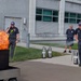 A fire safety training demonstration is taking place outdoors near an industrial or municipal building. A civilian in a blazer and khaki pants is being guided by a firefighter instructor on how to use a fire extinguisher on a controlled propane-fueled training fire. Several firefighters and onlookers are present, observing the exercise.