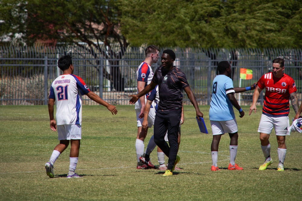 A soccer match is taking place on a grass field between two teams — the 'Spartans' in white/red/blue uniforms and a team in light blue uniforms. A coach or official in dark clothing holding a clipboard is seen interacting with players on the field. The scene appears to capture a mid-game moment, possibly a substitution or brief interaction near the sideline.