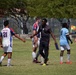 A soccer match is taking place on a grass field between two teams — the 'Spartans' in white/red/blue uniforms and a team in light blue uniforms. A coach or official in dark clothing holding a clipboard is seen interacting with players on the field. The scene appears to capture a mid-game moment, possibly a substitution or brief interaction near the sideline.