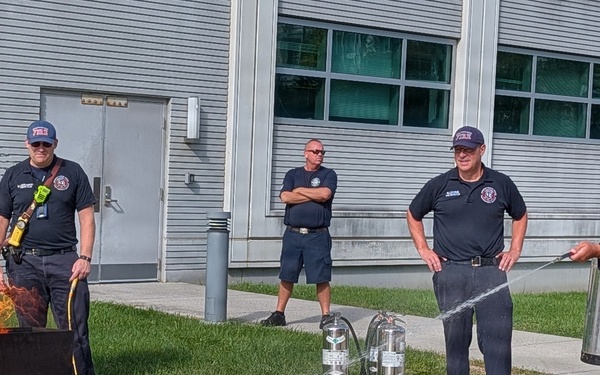 A fire safety training or demonstration is taking place outdoors near a building. A civilian in business casual attire is using a fire extinguisher on a controlled fire in a metal training pan, supervised by uniformed firefighters. Two additional firefighters observe in the background, with spare extinguishers staged on the ground nearby.