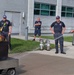 A fire safety training or demonstration is taking place outdoors near a building. A civilian in business casual attire is using a fire extinguisher on a controlled fire in a metal training pan, supervised by uniformed firefighters. Two additional firefighters observe in the background, with spare extinguishers staged on the ground nearby.