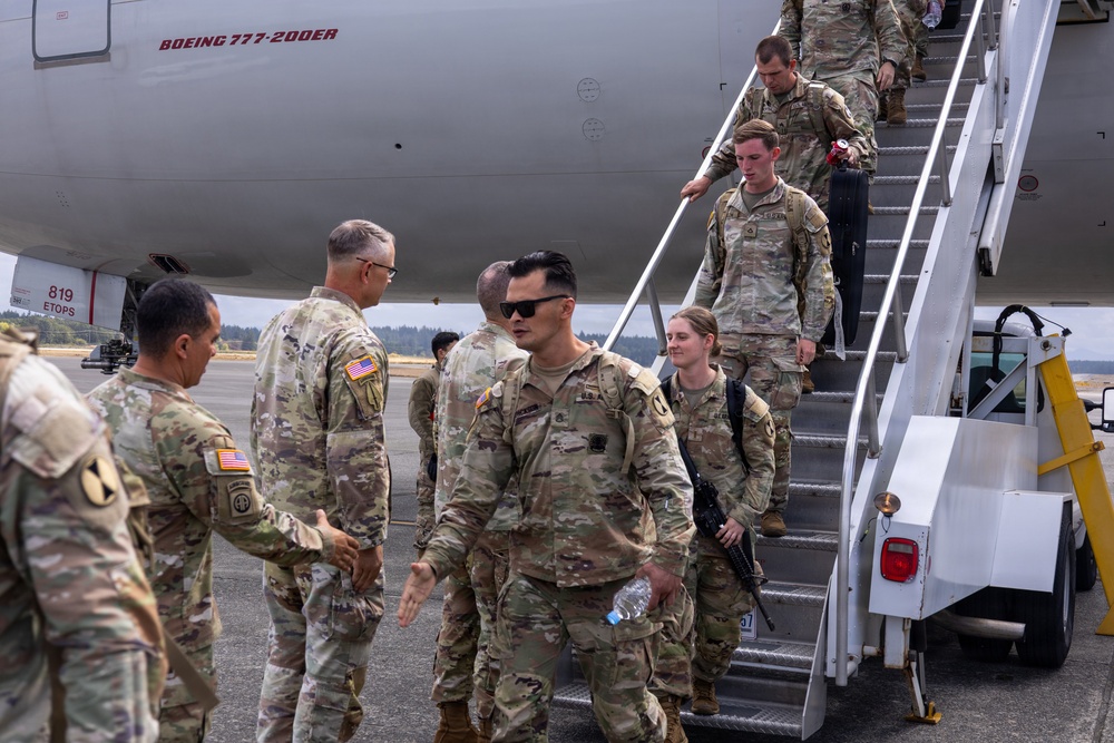 U.S. Army soldiers are deplaning from a Boeing 777-200ER aircraft via a mobile stairway onto a tarmac. Greeting personnel in uniform are shaking hands with arriving soldiers, suggesting a welcome-home or arrival ceremony. The setting appears to be a military airfield, likely in the Pacific Northwest based on the overcast sky and visible tree line.