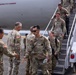 U.S. Army soldiers are deplaning from a Boeing 777-200ER aircraft via a mobile stairway onto a tarmac. Greeting personnel in uniform are shaking hands with arriving soldiers, suggesting a welcome-home or arrival ceremony. The setting appears to be a military airfield, likely in the Pacific Northwest based on the overcast sky and visible tree line.