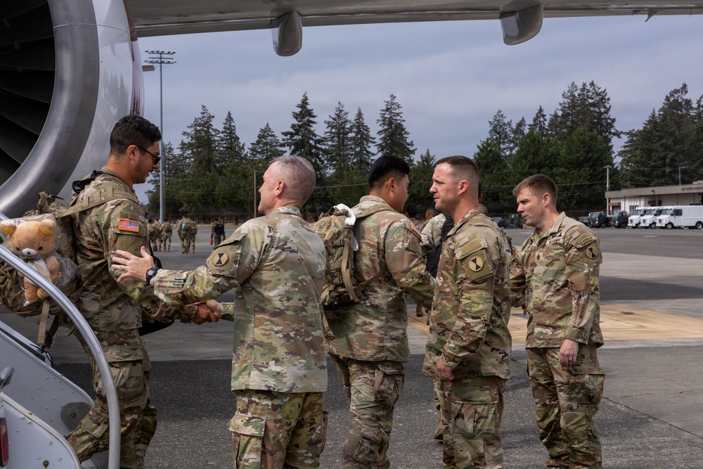 A group of U.S. Army soldiers in OCP (Operational Camouflage Pattern) uniforms are gathered on an airfield tarmac next to a large jet aircraft engine. A handshake is taking place between two soldiers, suggesting a greeting or farewell ceremony. The setting appears to be a military airfield with trees, light poles, and vehicles visible in the background.
