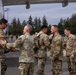 A group of U.S. Army soldiers in OCP (Operational Camouflage Pattern) uniforms are gathered on an airfield tarmac next to a large jet aircraft engine. A handshake is taking place between two soldiers, suggesting a greeting or farewell ceremony. The setting appears to be a military airfield with trees, light poles, and vehicles visible in the background.