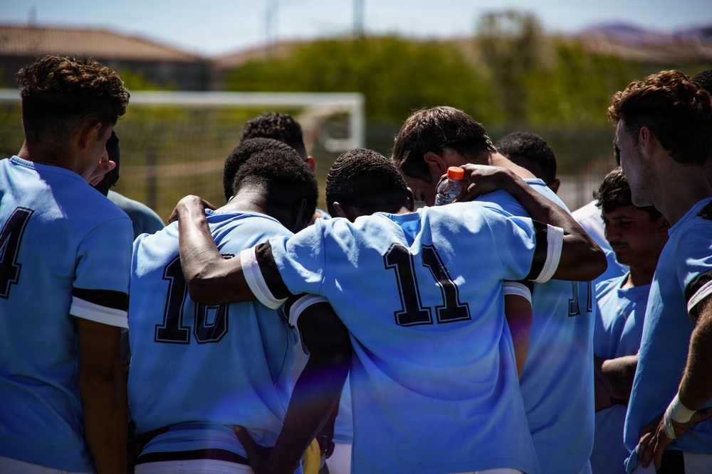 A soccer team dressed in light blue and dark-trimmed jerseys forms a tight huddle on an outdoor field, with players placing arms around each other's shoulders. The scene appears to be either a pre-game or post-game team gathering. A soccer goal post is visible in the background along with hills and a clear sky.