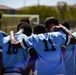 A soccer team dressed in light blue and dark-trimmed jerseys forms a tight huddle on an outdoor field, with players placing arms around each other's shoulders. The scene appears to be either a pre-game or post-game team gathering. A soccer goal post is visible in the background along with hills and a clear sky.