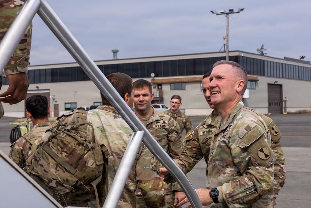 A group of U.S. Army soldiers in Operational Camouflage Pattern (OCP) uniforms are gathered on what appears to be a military installation tarmac or airfield. A senior soldier with the name tape 'RICHTER' is prominently visible on the right, appearing to engage in conversation with fellow soldiers. The setting suggests an arrival, departure, or greeting ceremony near a military building.