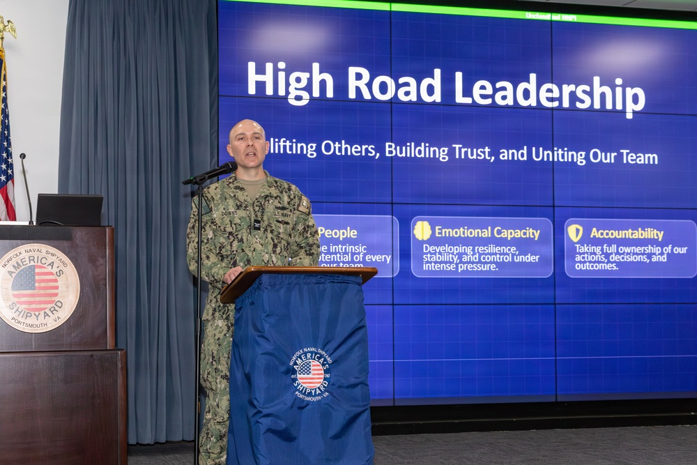 A U.S. Navy service member identified by nametape as 'DOWNS' is presenting at a podium at Norfolk Naval Shipyard in Portsmouth, VA. The presentation is titled 'High Road Leadership' with the subtitle 'Uplifting Others, Building Trust, and Uniting Our Team.' The slide displays three key concepts: People, Emotional Capacity, and Accountability.