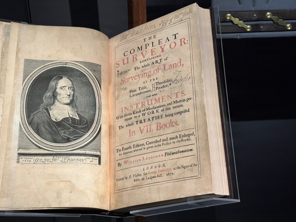 An open antique book displayed on a stand, showing a title page and an engraved portrait frontispiece. The book is 'The Compleat Surveyor' by William Leybourn, fourth edition, printed in London in 1679. The title page features red and black typography typical of 17th-century printing, and the left page contains an engraved oval portrait of the author.